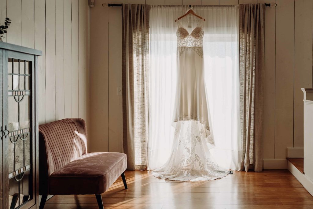 wedding dress hanging in the a window backlit by the sun in a entry way at Stonehurst at Hampton Valley Connecticut wedding venue by Ladman Studios