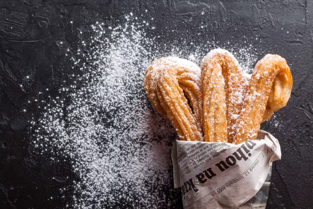 A plate of freshly fried churros with a powdered sugar on the side wrapped in a new print napkin.