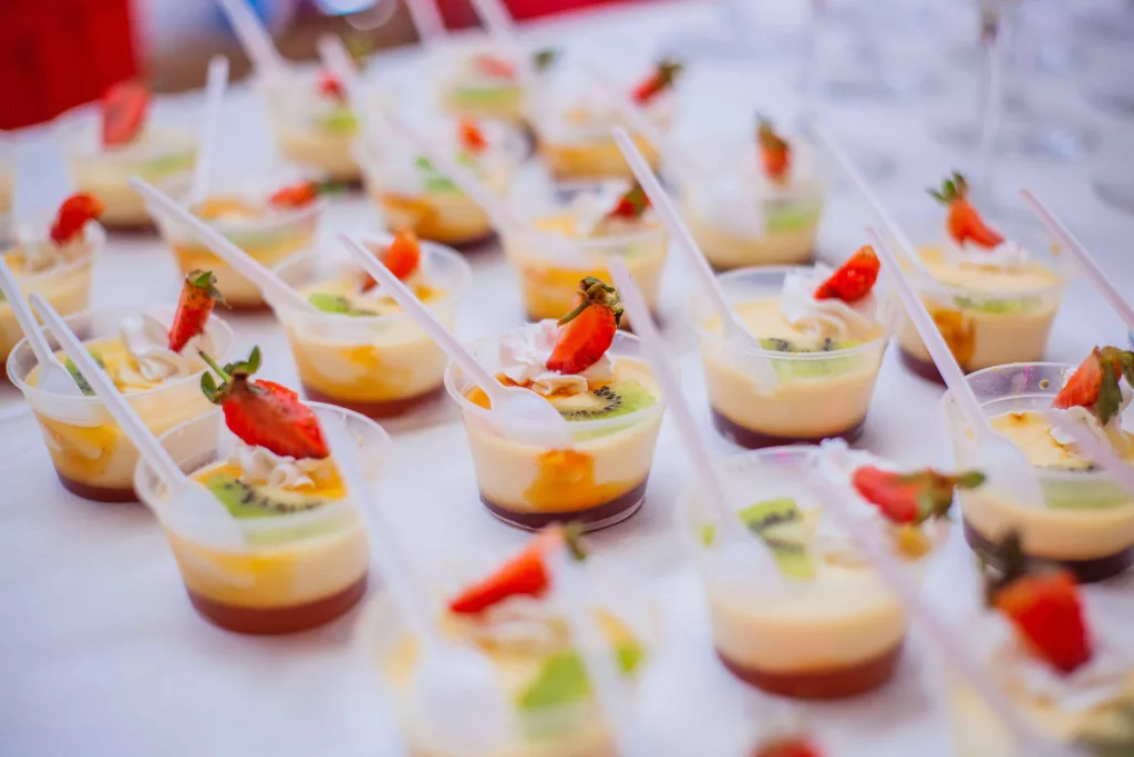 A tray of small dessert shots arranged in rows, with colorful layers of cream, fruit, and cake visible through the clear glass. Each shot is garnished with a fresh berry or sprinkle of chocolate shavings. The tray sits on a wooden table with a white tablecloth and a few decorative flowers in the background.