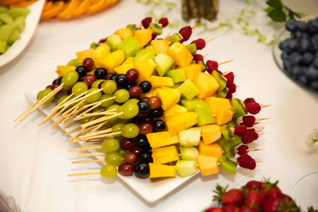 A plate of colorful fruit skewers featuring chunks of fresh pineapple, juicy strawberries, sweet grapes, and slices of tangy kiwi. The skewers are neatly arranged on a white platter, with a sprinkle of powdered sugar adding a touch of sweetness to the fruity goodness. The perfect snack for a hot summer day!