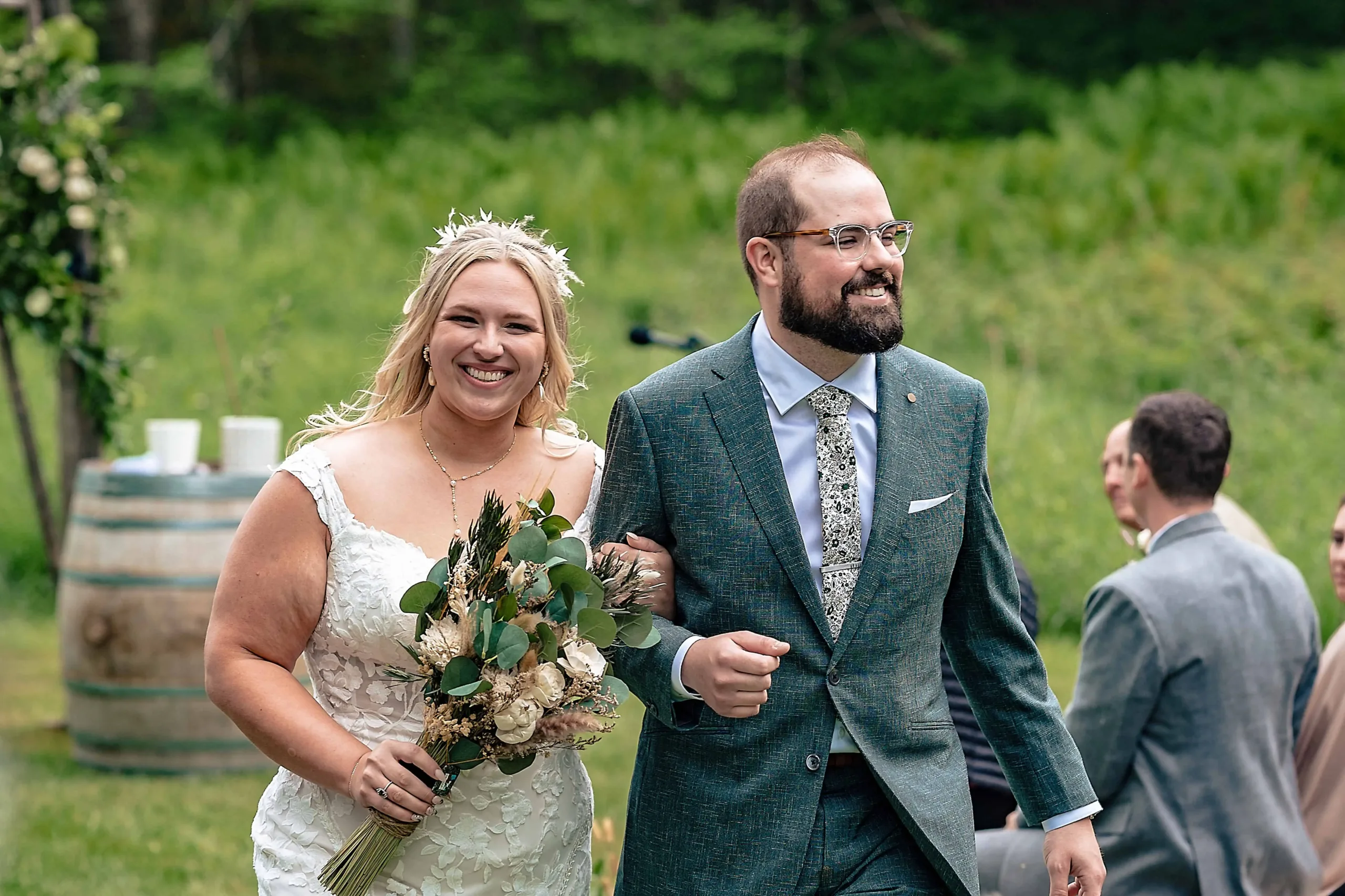 A smiling couple in wedding attire walk together outdoors in Connecticut; the bride in a lace dress with a floral bouquet and the groom in a green tweed suit.
