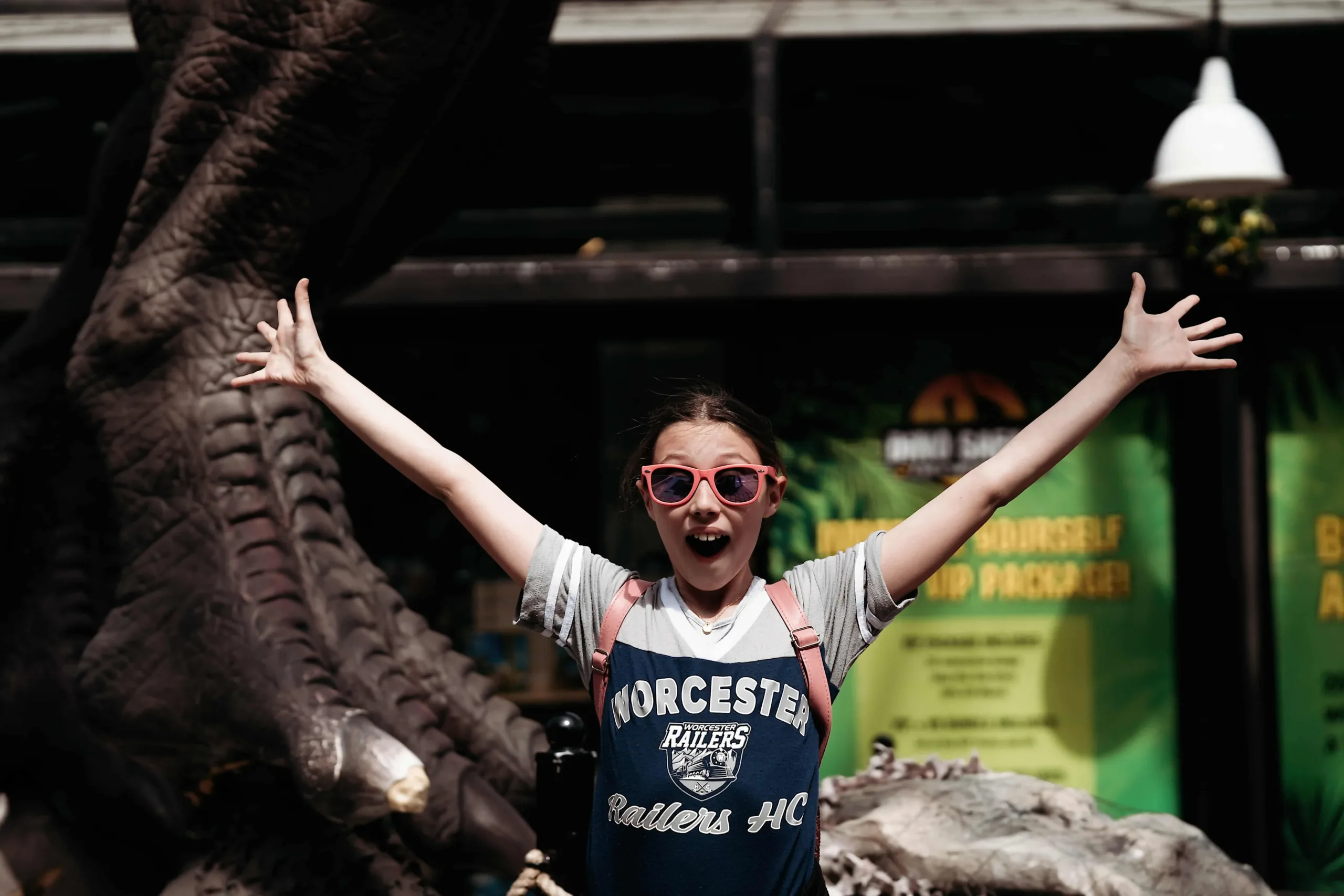 A delighted girl with arms raised in front of a dinosaur exhibit, capturing the spontaneous moments that make documentary family photography essential.