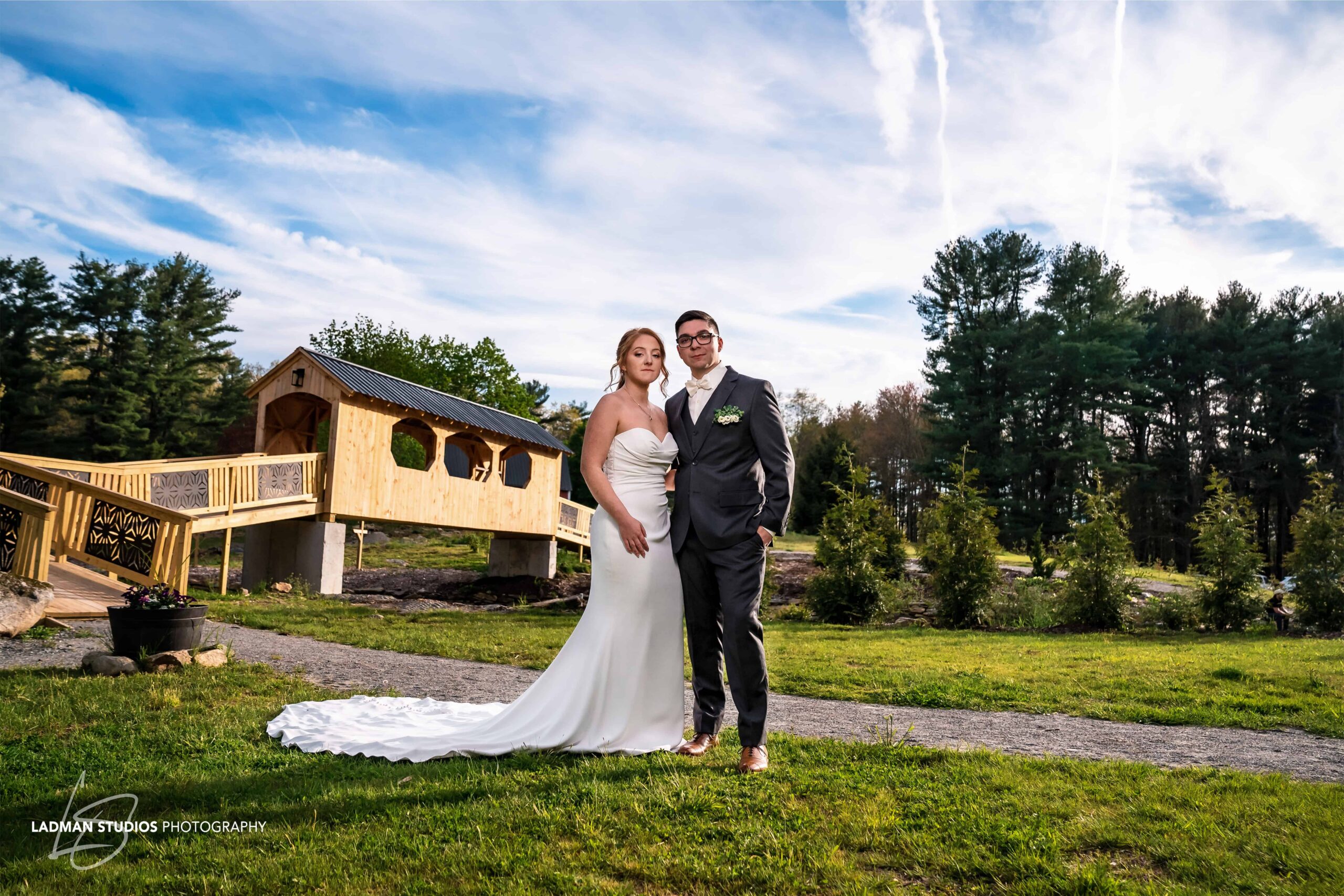 A bride and groom standing in front of a wooden bridge in a rural setting, with trees and a clear sky in the background.