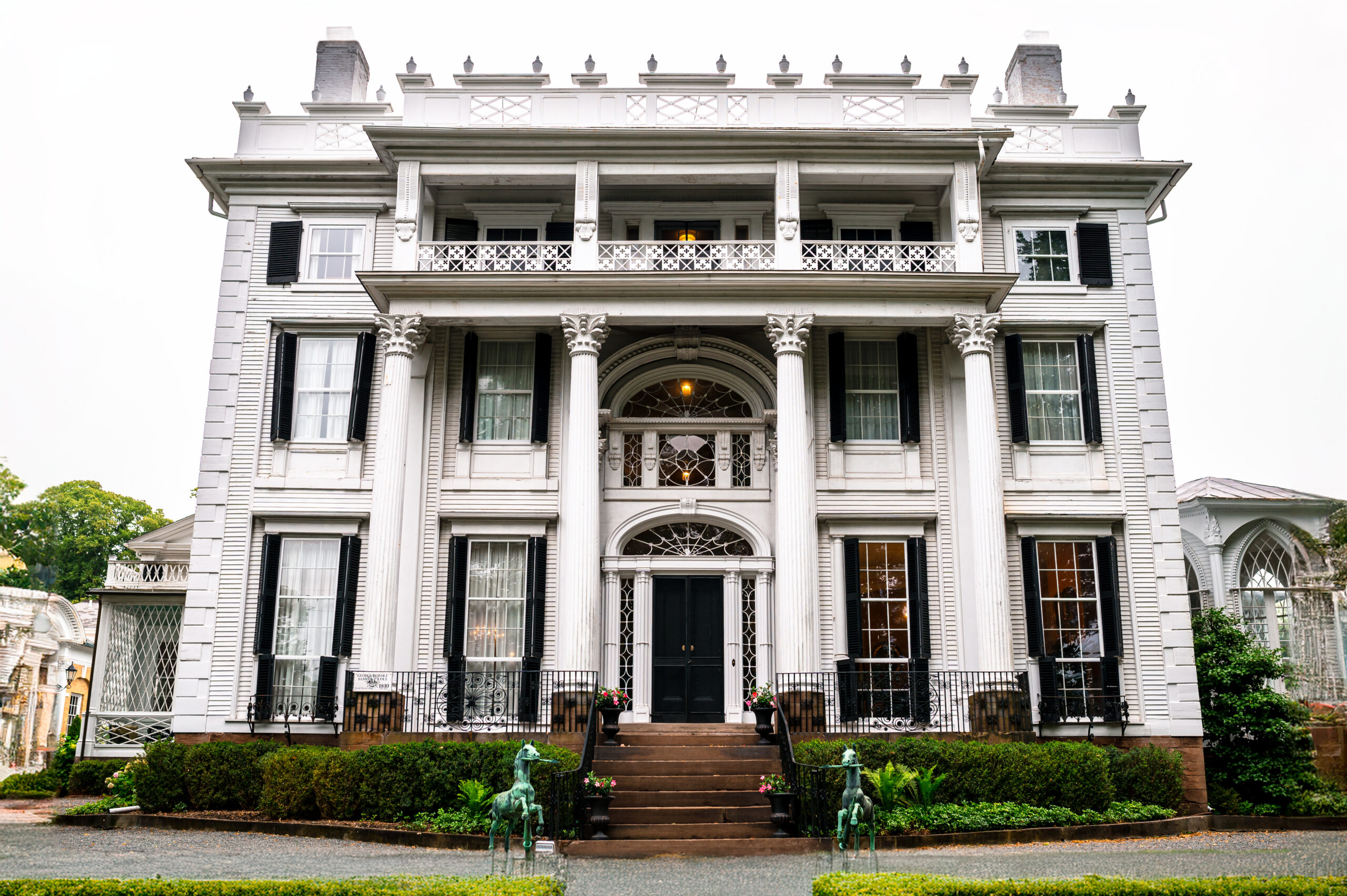 Linden Place, a stately white mansion with grand Ionic columns, intricate black window shutters, and a decorative wrought iron balcony, surrounded by lush gardens and statues