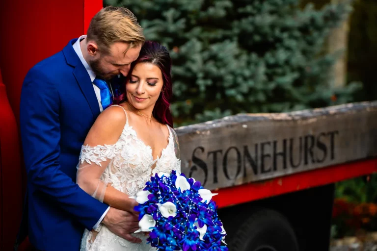 A groom in a blue suit tenderly embraces his bride, dressed in an intricate lace wedding gown, as she holds a vibrant bouquet of blue and white flowers, taken by a Connecticut wedding photographer