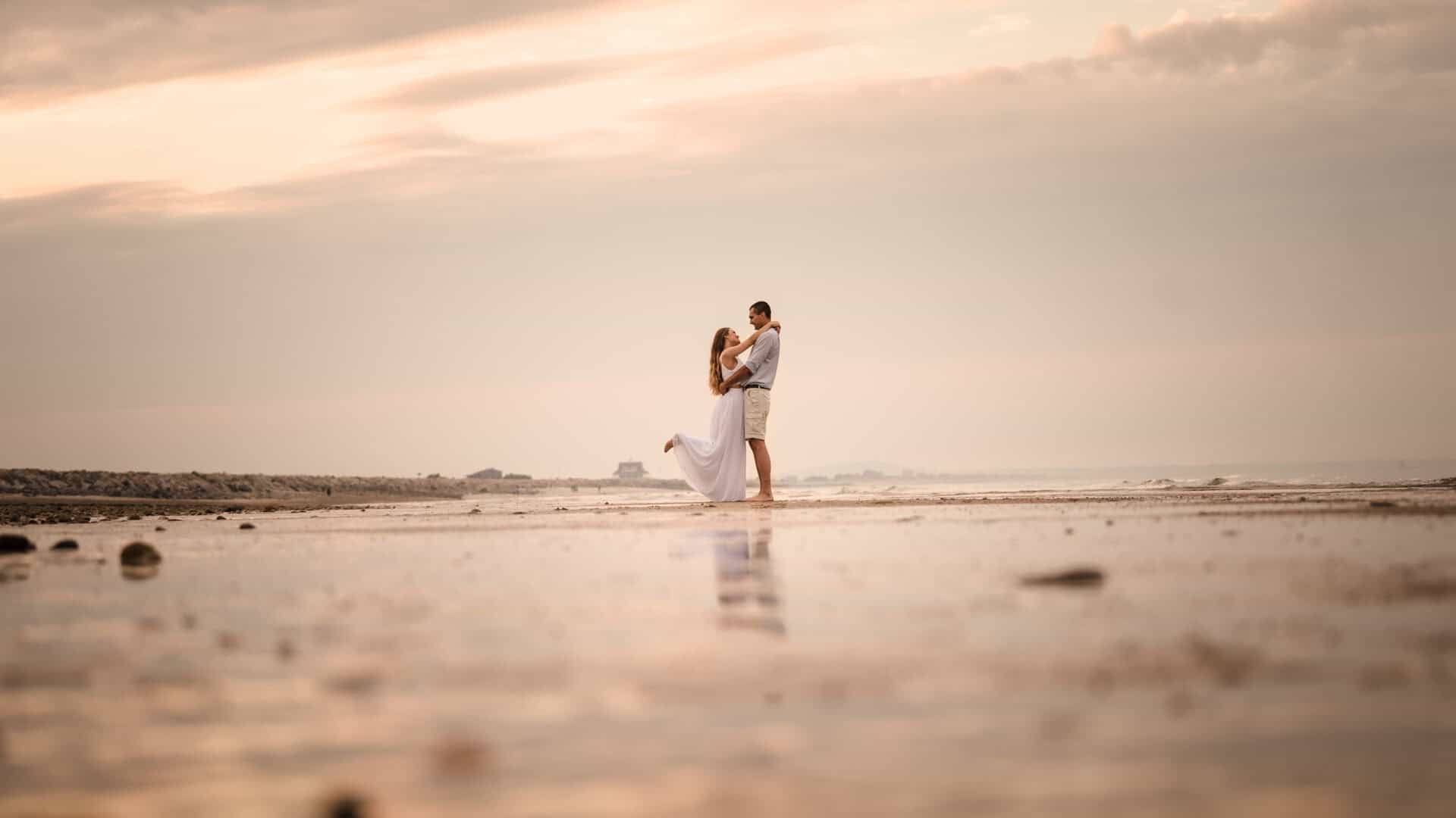 A couple embraces on a serene beach at sunset, with soft pastel skies and reflections on the wet sand, captured by a Connecticut engagement photographer.