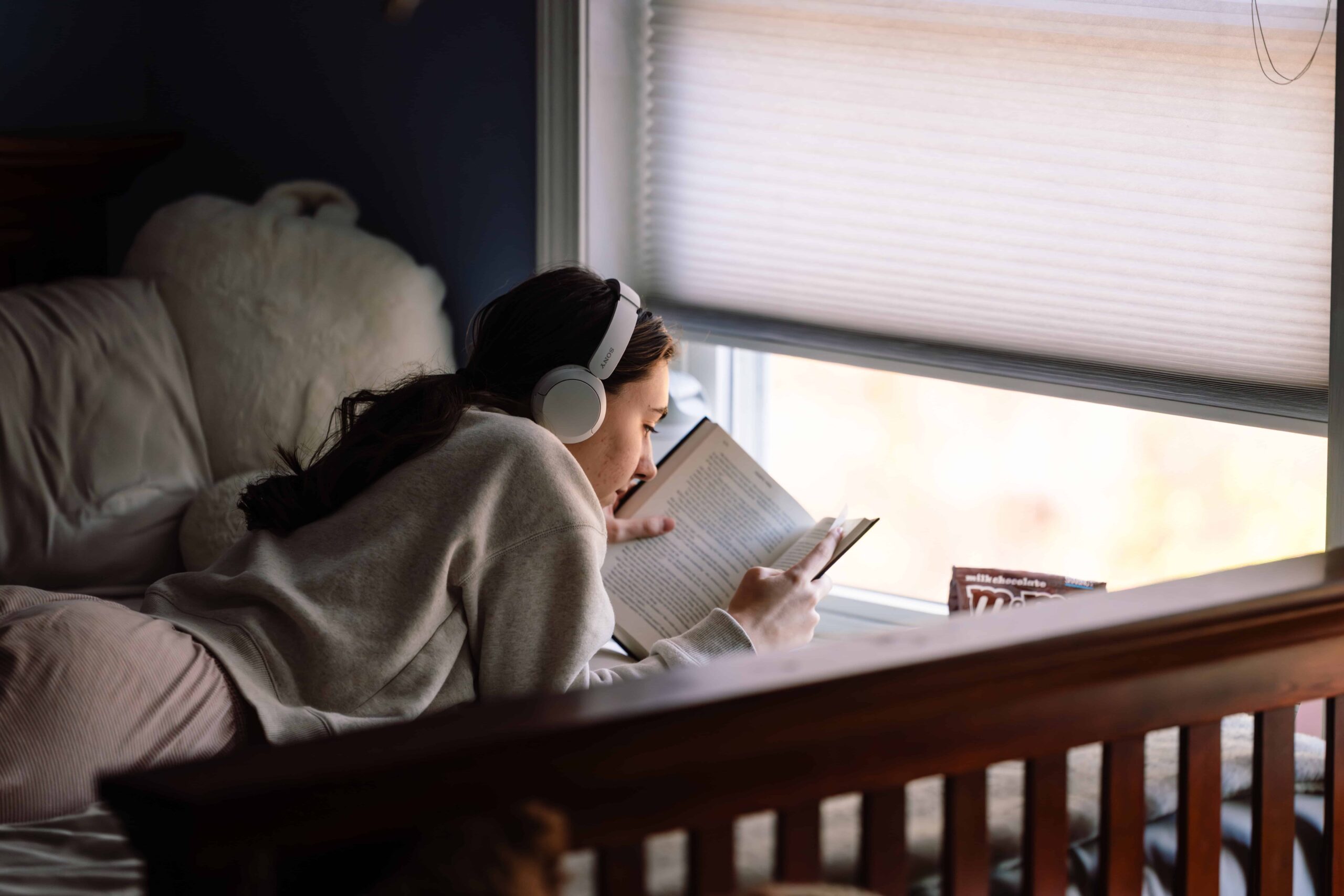 A teenager reading by the window with headphones on during a quiet moment at home, photographed by a Connecticut documentary family photographer.